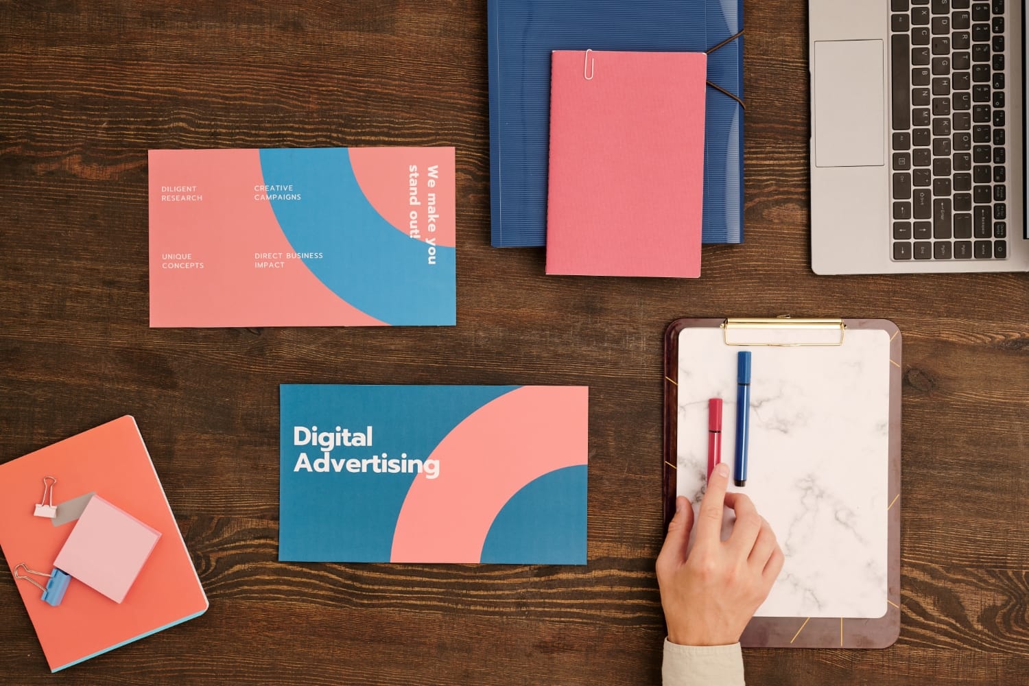 Hand arranging colorful marketing materials, including a brochure labeled "Digital Advertising" and a card stating "We make you stand out," on a wooden desk, reflecting graphic design and branding services.