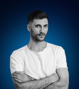 Man with crossed arms wearing a white t-shirt against a blue background, representing a confident professional in digital marketing and web design.